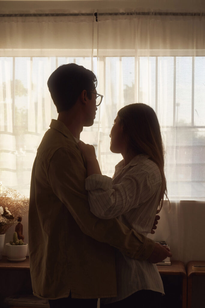 Gaby and Me a couple stand close by a window bathed in soft sunlight sharing an intimate moment.